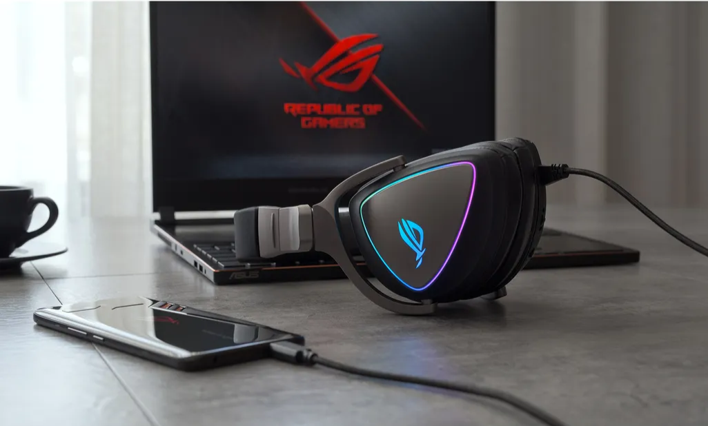 Gaming headset with RGB lighting in front of a laptop displaying the ASUS ROG logo.
